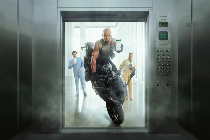 A man rides a motorcycle inside an elevator, holding up a mobile phone, with two other people standing in the elevator's background.